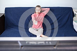 Pre-teen age girl watching television, sits on couch with tv remote control, wide screen is on foreground