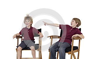 Pre-teen boy pointing at friend's hair while sitting on director's chair over white background