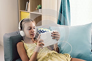 Pre teen child playing tablet and listening to music in head phones while relaxing on couch in living room at home