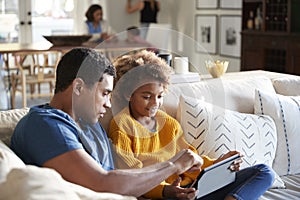 Pre-teen girl sitting on sofa in the living room using tablet computer with her father, mother and toddler sitting at a table in t