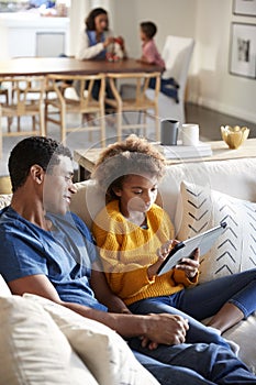 Pre-teen girl sitting on sofa in the living room using tablet computer with her father, mother and toddler sitting at a table in t