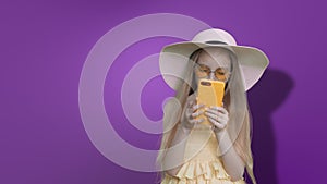 Pre-teen girl in summer hat reads phone on lilac wall background, alpha channel.