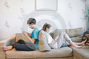 Pre-teen sister and brother gaming online with cellphones while sitting at home, children wearing safety masks, sitting back to