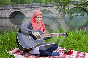 Preaty muslim girl playing acoustic guitar on the river bank