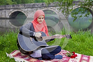 Preaty muslim girl playing acoustic guitar on the river bank