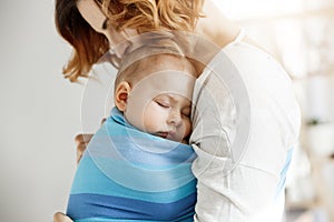 Precious little newborn boy having deep sleep at day on mother chest in blue baby sling. Mom kissing baby head and