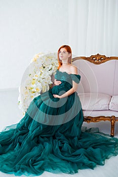 Pregnancy beauty. Beautiful elegant readhead pregnant woman in green dress posing in tender home interior.