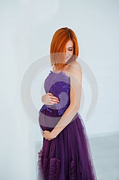 Pregnancy beauty. Beautiful elegant readhead pregnant woman in violet dress posing in tender home interior.