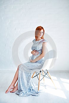Pregnancy beauty. Beautiful elegant readhead pregnant woman in white dress posing in tender home interior.