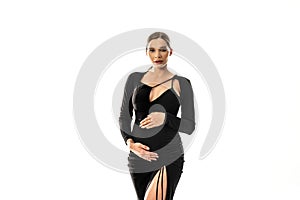 Pregnancy concept. Woman posing in white studio background wearing black dress, holding her pregnant belly, looking at the camera