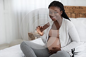 Pregnancy Cravings. Smiling Expectant Black Woman Eating Milk Chocolate Bar At Home