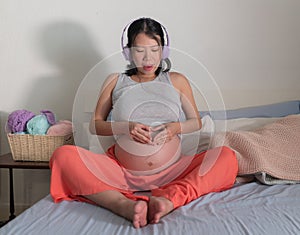 Pregnancy lifestyle home portrait of young happy and beautiful Asian Japanese woman sitting pregnant on bed listening to music