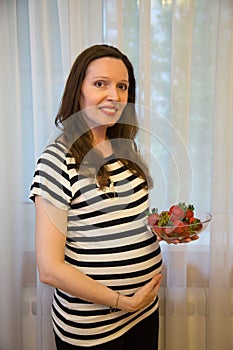 Pregnancy, motherhood, the concept of waiting-close - up of a happy pregnant woman with a big belly in a window with strawberries