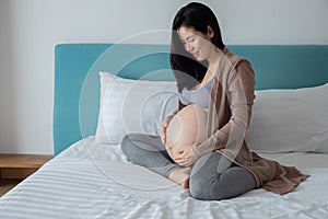 Pregnancy, motherhood and expectation concept - smiling asian  pregnant woman touching big belly on bed