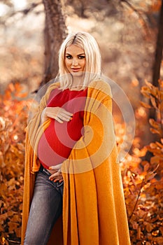 Pregnancy. Motherhood. Pregnant woman hugging her belly enjoying in autumn forest park. Beautiful blond woman in red knit sweater