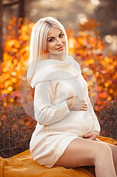 Pregnancy. Motherhood. Pregnant woman hugging her belly enjoying in autumn forest park. Beautiful blond woman in white knit