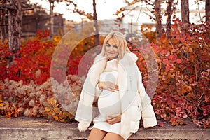 Pregnancy. Motherhood. Pregnant woman hugging her belly enjoying in autumn forest park. Beautiful blond woman in white knit