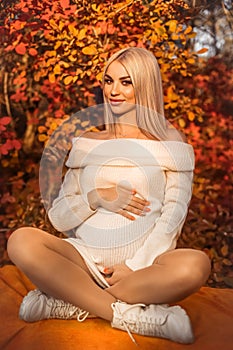 Pregnancy. Motherhood. Pregnant woman hugging her belly enjoying in autumn forest park. Beautiful blond woman in white knit