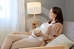 Pregnancy nourishment joy. Relaxed pregnant woman touching her belly sitting on sofa holding bowl with fresh fruit looking at