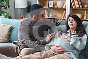 Pregnancy and people concept - happy African man hugging pregnant woman at home. Young couple sitting on sofa, husband support and