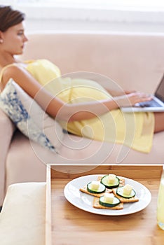 Pregnancy platter. a pregnant woman using a laptop at home with a healthy meal of crackers and cucumbers next to her.