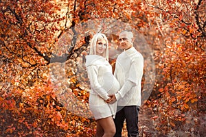 Pregnancy. Pregnant couple in white outfits enjoying in autumn forest park. Beautiful blond woman with husband. Parenthood