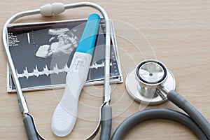 Pregnancy test showing a positive result and stethoscope, ultrasound picture of baby  on wooden background. Result of ultr