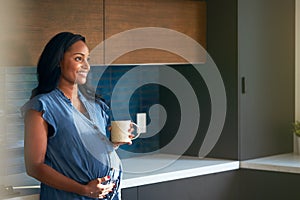 Pregnant African American Woman Standing In Kitchen At Home With Hot Drink