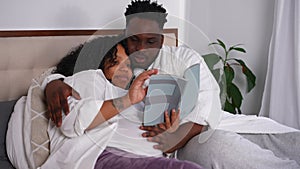 Pregnant African American young woman reading book lying in bed with man talking. Happy relaxed couple enjoying leisure