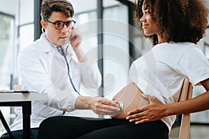 Pregnant african woman has appointment with doctor at clinic. Male gynaecologist OB GYN medic specialist with stethoscope listens