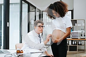 Pregnant african woman has appointment with doctor at clinic. Male gynaecologist OB GYN medic specialist with stethoscope listens