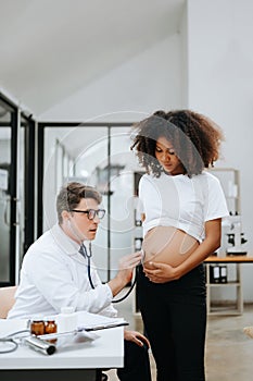 Pregnant african woman has appointment with doctor at clinic. Male gynaecologist OB GYN medic specialist with stethoscope listens