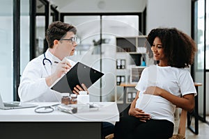 Pregnant african woman has appointment with doctor at clinic. Male gynaecologist OB GYN medic specialist with stethoscope listens photo