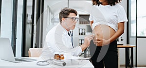 Pregnant african woman has appointment with doctor at clinic. Male gynaecologist OB GYN medic specialist with stethoscope listens photo