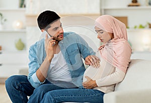 Pregnant arab woman having prenatal contractions, worried husband calling doctor, using mobile phone, home interior