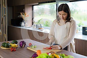 Pregnant Asian woman cutting tomato for fresh green salad, female prepares tasty organic dinner at home, healthy nutrition for