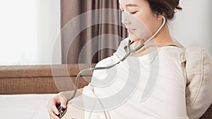 Pregnant Asian woman using stethoscope listening to baby heartbeat in white bedroom.