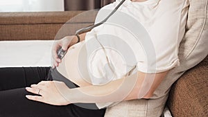 Pregnant Asian woman using stethoscope listening to baby heartbeat in white bedroom.