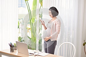 Pregnant asian woman working from home in modern office contacting using mobile phone in call talking