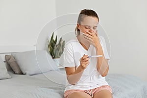 Pregnant attractive woman wearing casual attire sitting on sofa at home and holding pregnancy test with positive result, suffering