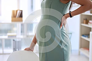 Pregnant, back pain and woman in office and standing for relief, stretching and comfort by chair. Maternity, corporate