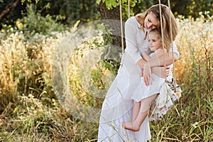 Pregnant beautiful mother with little blonde girl in a white dress sitting on a swing, laughing, childhood, relaxation