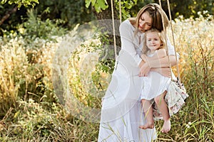 Pregnant beautiful mother with little blonde girl in a white dress sitting on a swing, laughing, childhood, relaxation