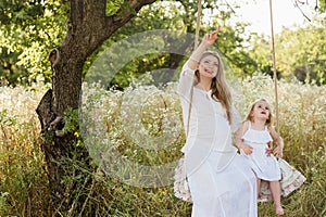 Pregnant beautiful mother with little blonde girl in a white dress sitting on a swing, laughing, childhood, relaxation, serenity,