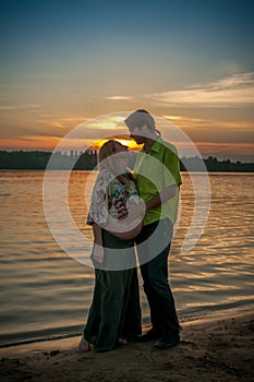 A pregnant beautiful woman with her husband on the river bank beach smiling and touching her belly with love and care