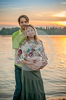 A pregnant beautiful woman with her husband on the river bank beach smiling and touching her belly with love and care
