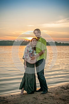 A pregnant beautiful woman with her husband on the river bank beach smiling and touching her belly with love and care