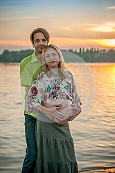 A pregnant beautiful woman with her husband on the river bank beach smiling and touching her belly with love and care