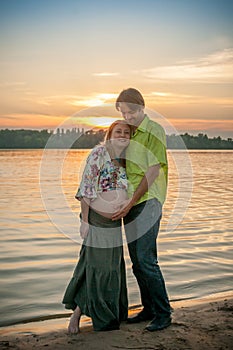 A pregnant beautiful woman with her husband on the river bank beach smiling and touching her belly with love and care. Happy coupl