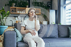 Pregnant beautiful woman sitting on the couch at home, talking on a cell phone with a doctor, calling an ambulance, giving birth